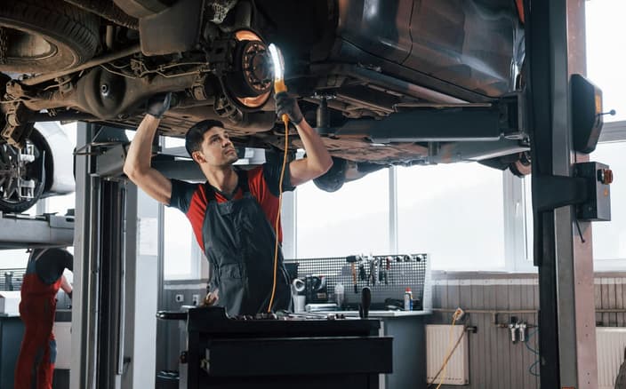 Mechanik v servisu provádí diagnostiku podvozku, se světlem kontroluje tyčky stabilizátoru a nápravu na zvednutém autě.