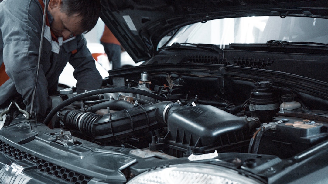 Mechanik v šedém pracovním oblečení se sklání nad otevřeným motorovým prostorem moderního vozu v dílně a kontroluje motor. V popředí je skříň vzduchového filtru a sací potrubí.