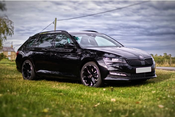 Черная Шкода Суперб Комби (Škoda Superb III) на фоне загородного пейзажа и облачного неба с оригинальными черными дисками.