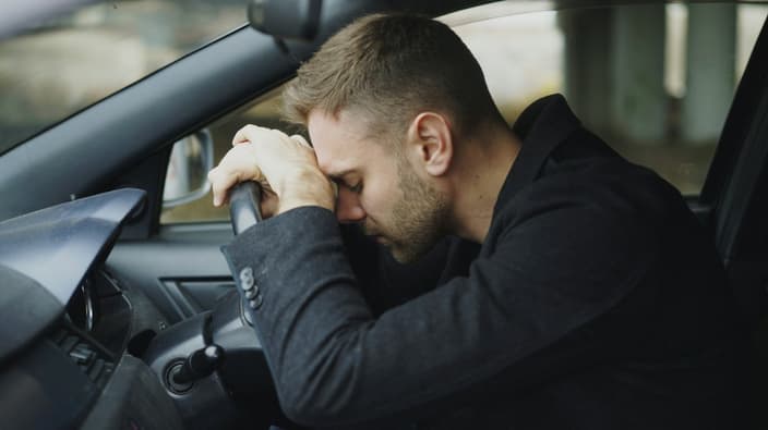 Stresovaný muž opírá hlavu o volant, frustrovaný z poruchy auta a potřeby urgentní opravy v autoservisu.