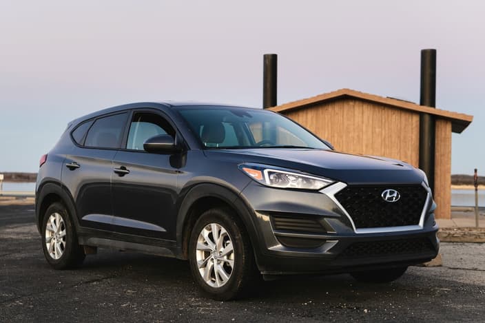 Tmavě šedé SUV Hyundai Tucson zaparkované na asfaltové ploše u dřevěné budovy během západu slunce.