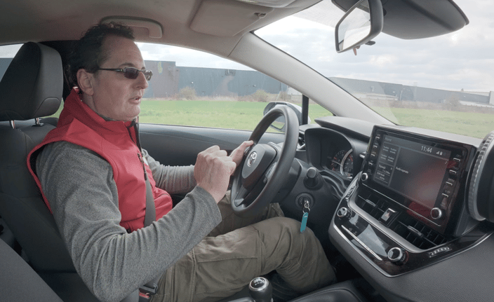 Muž v červené vestě za volantem Toyoty Corolly, soustředěný na řízení, s viditelným interiérem vozu včetně palubní desky a multimediálního displeje.