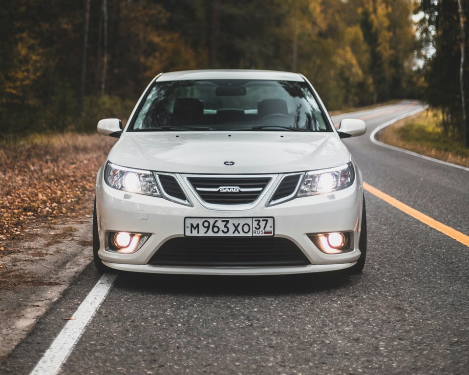 Klasický bílý sedan Saab 9-3 na malebné silnici, což naznačuje náš specializovaný servis pro náročné majitele automobilů a unikátní značky.