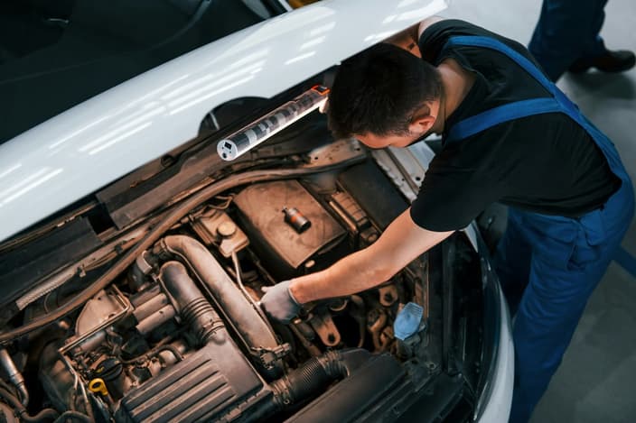 Mechanik s baterkou kontroluje motor auta, zaměřuje se na alternátor a startér pro diagnostiku problémů s elektřinou.
