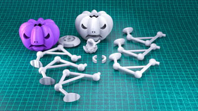 A still life showing various 3D printed objects on a turquoise cutting mat with a grid. The objects include two pumpkin-shaped figures (one purple, one white/grey) and several white/grey interconnected modular plastic parts around them.