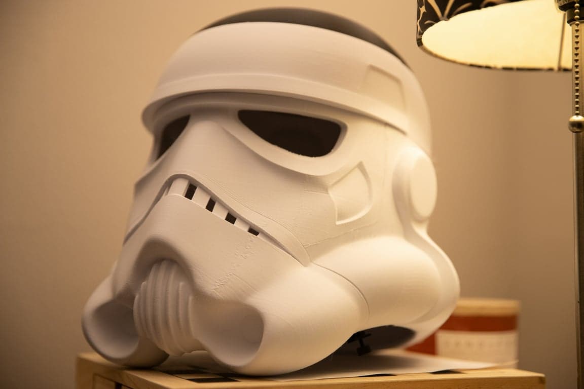A 3D printed Stormtrooper helmet, in an unfinished matte white state with visible layer lines from the printing process, rests on a wooden surface next to a lamp, showcasing a work-in-progress cosplay prop.