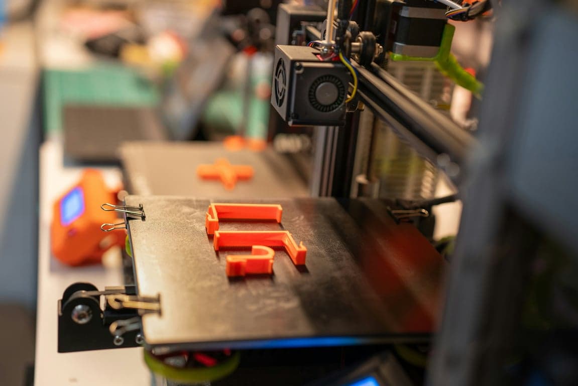 In a shallow depth-of-field photograph, a black 3D printer build plate features a partially printed orange 'F' shape and an orange plus shape, secured by metal clips. In the out-of-focus background, other printer elements are visible, including a fan and electronic components. The ambient lighting suggests an indoor setting, such as a workshop or office.